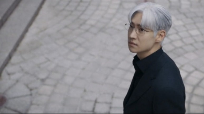 검은 정장을 입은 남성이 거리에서 의미심장한 표정으로 뒤를 돌아보는 장면