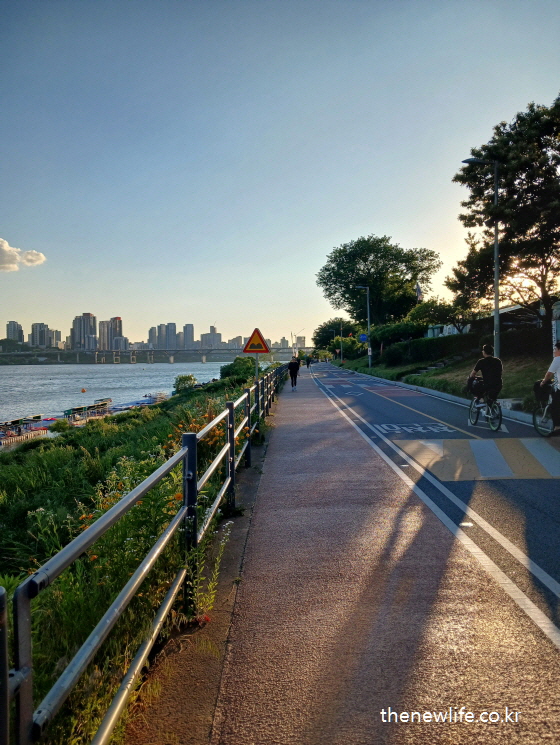 Sunset along the Han River with people walking and cycling, the skyline visible in the distance./해 질 무렵 한강 자전거 도로에서 운동 중인 사람들과 멀리 보이는 도시 스카이라인.