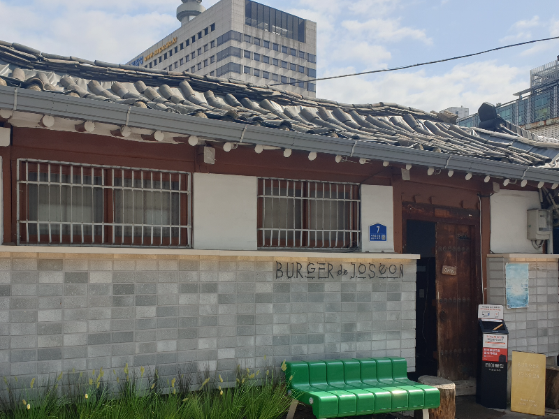 기와 지붕에 나무 대문, 그리고 요즘 벽돌로 쌓은 담. 그 앞에는 초록색 벤치가 있다.