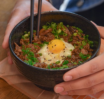 동그란 검은 그릇에 소고기 볶은 것을 밥 위에 올리고 가운데 살짝 익힌 계란을 두었다. 잘게 썬 파로 장식했고 검은 깨를 뿌려 먹음직스럽게 장식한 소고기 달걀 덮밥.