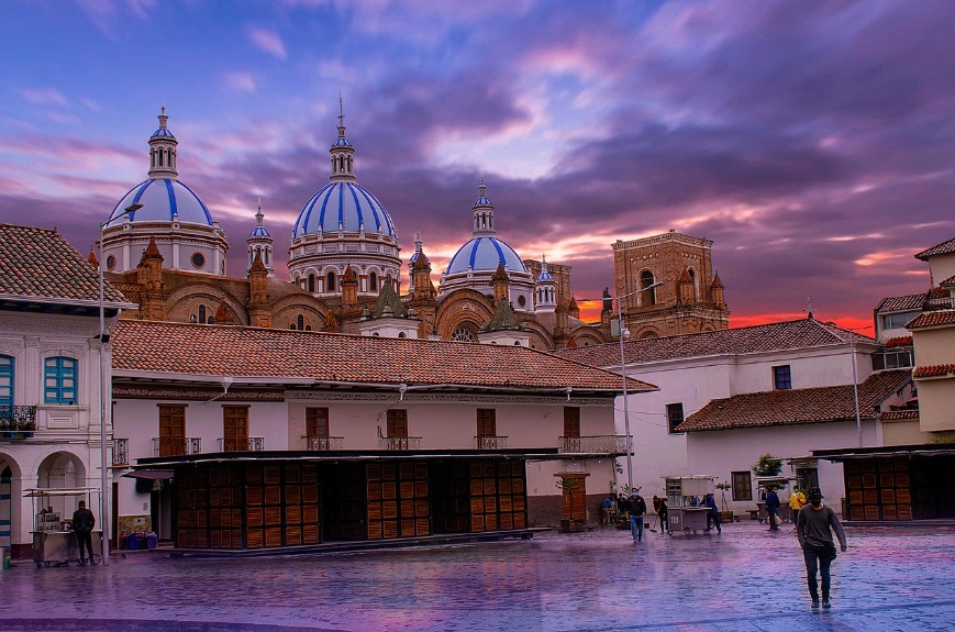노을이 지는 보랏빛 하늘을 배경으로 파란색 돔과 고풍스러운 석조 건축물이 웅장하게 서 있는 에콰도르 쿠엥카의 뉴 카테드랄 대성당과 그 앞 광장의 전경입니다.