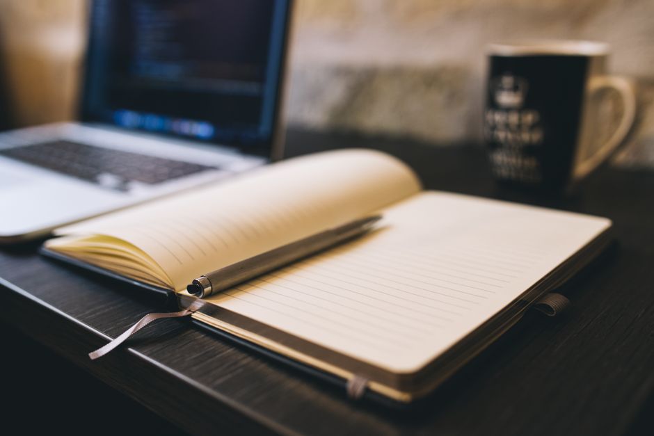 Open notebook with a pen on a wooden desk beside a laptop, ideal for blogging and work-related themes.