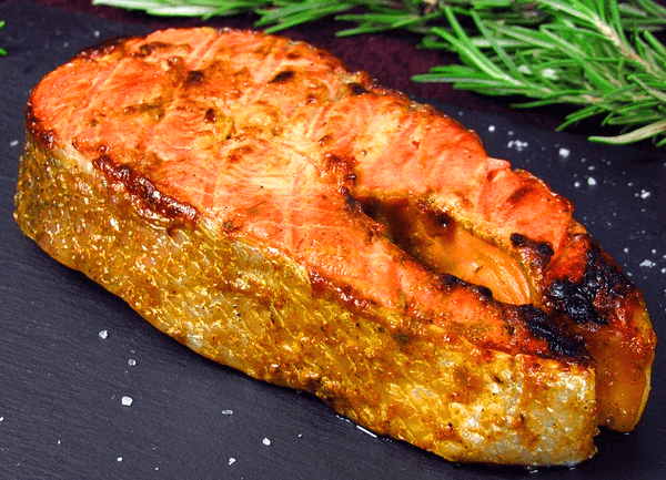 검정 접시 위체 두툼한 연어스테이크 한 조각이 있고, 하얀 소금이 뿌려진 상태다. 오른 쪽에는 초록색 로즈마리가 있다.