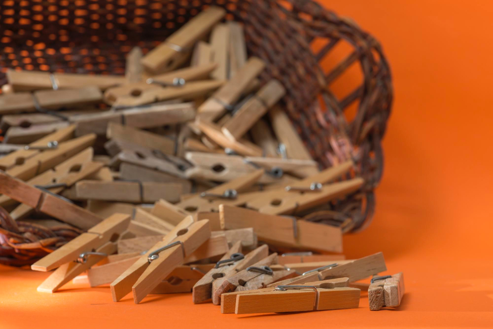 오렌지색 배경에 갈색 바구니에서 쏟아져 나온 나무 빨래집게들. 전통적인 베이지색 목재 빨래집게가 바구니 밖으로 흩어져 있는 따뜻한 분위기의 생활용품 이미지