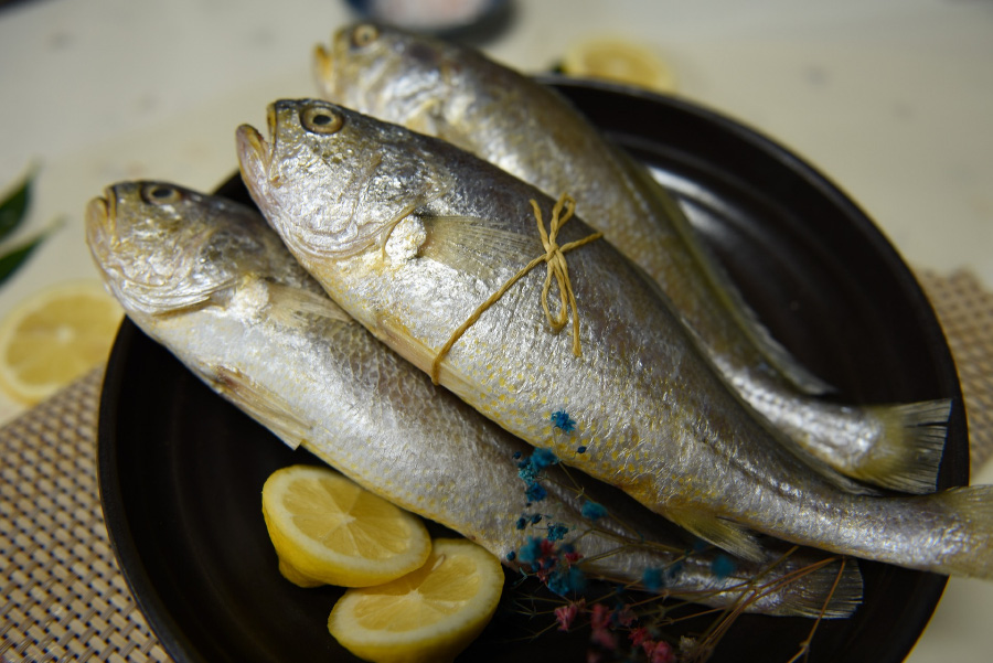 바다에서 채취를 한 조기를 잘 손질을 한 후에 구운 것을 검은 색의 동그란 접시 위에 3마리를 레몬 조각과 꽃나무와 함께 놓아두고 찍은 사진