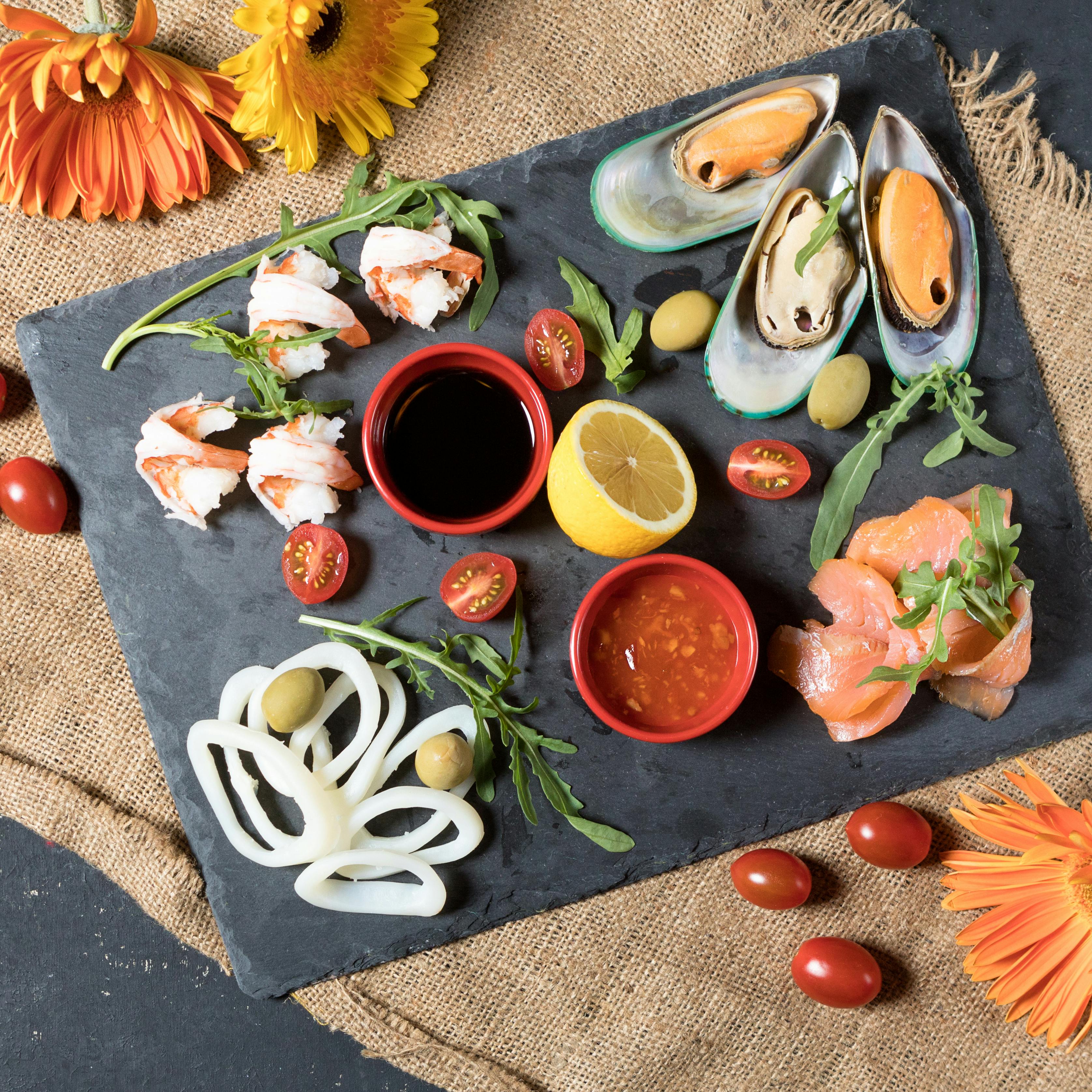 A flat-lay view of a seafood platter on a dark slate surface, featuring mussels, shrimp, salmon, squid, tomatoes, and dipping sauces, garnished with arugula and surrounded by burlap and vibrant orange gerbera daisies.