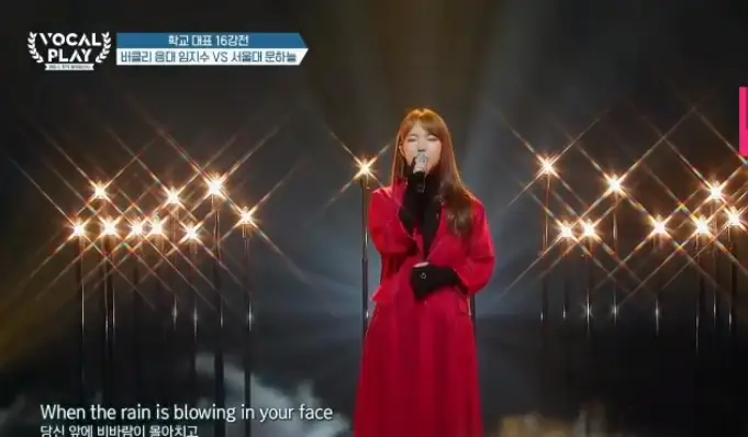 싱어게인3- 무대위 빨간 롱코트를 입고 갈색긴머리를 늘어뜨린채 오른손으로 마이크를 들고 눈감고 노래하는 27호