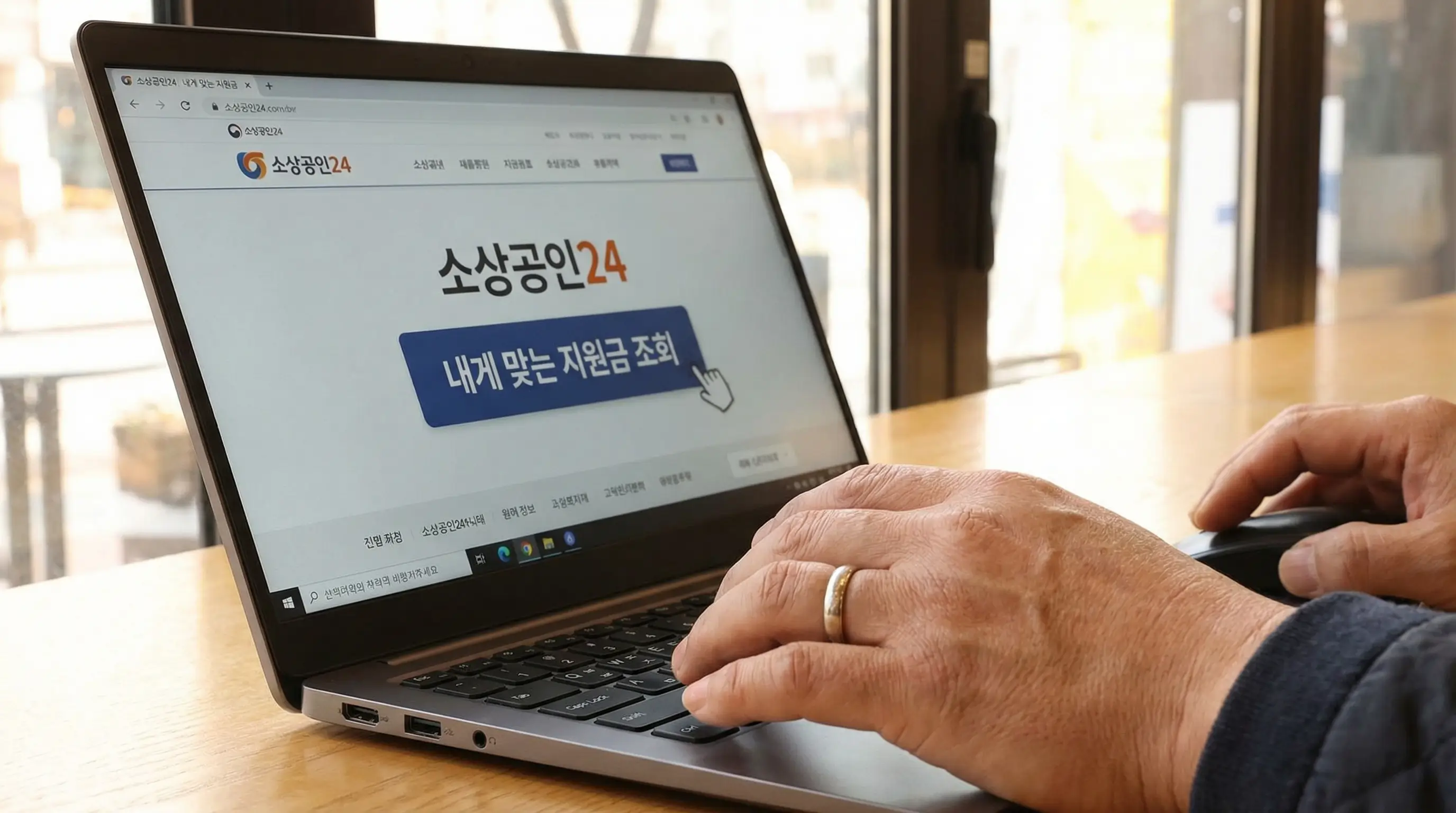 노트북 화면에 소상공인24 홈페이지를 띄워놓고 내게 맞는 지원금을 조회하는 사장님의 손 클로즈업