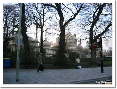 영국 브라이턴에 위치한 로열 파빌리온이 나무 사이로 보이는 거리 풍경. 이국적인 인도-이슬람 양식의 돔과 첨탑이 인상적이며, 인도를 걷는 사람들과 버스 정류장 정보판이 도시의 일상적인 분위기를 더해준다.