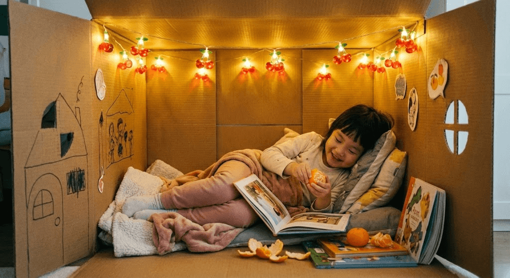 앵두 전구로 장식된 아늑한 종이 박스 집 안에서 귤을 먹으며 책을 보는 아이