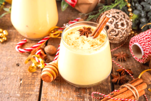 손잡이가 있는 따뜻한 머그잔에 담긴 크림색 에그녹 칵테일. 넛맥 가루가 뿌려져 있고, 잔 옆에 진저 브레드 쿠키가 놓여 있어 아늑하고 달콤한 느낌을 강조합니다.