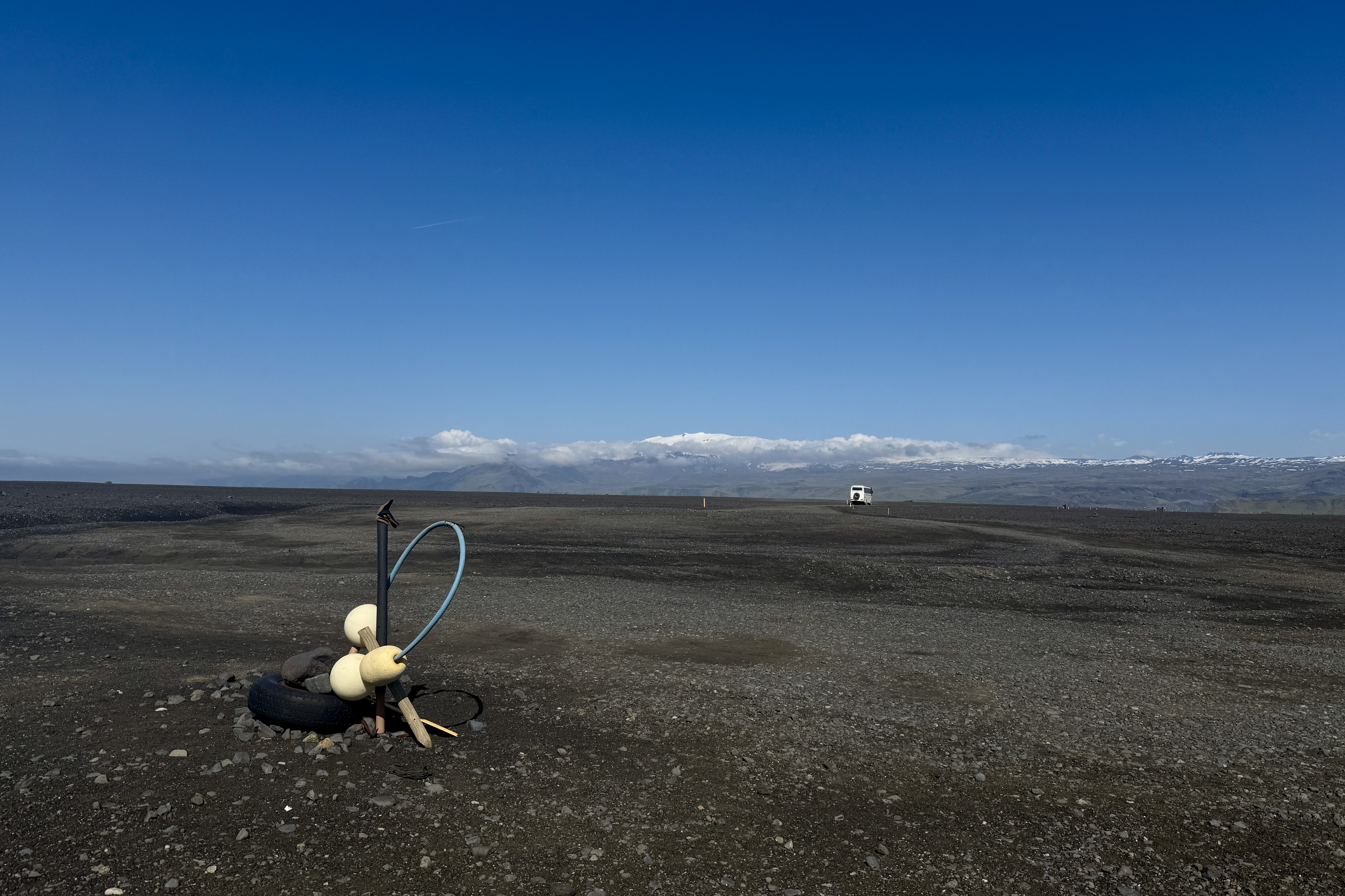광활한 검은 화산재 평원의 모습으로, 먼 거리에 하얀 차량이 보이고 전경에는 타이어와 막대로 만든 작은 구조물이 있습니다. 배경에는 맑은 푸른 하늘과 눈 덮인 산맥이 보입니다.