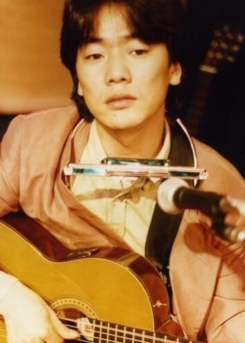 김광석이-기타를-메고-목에는-하모니카를-걸고-어딘가를-응시하고-있는-사진
