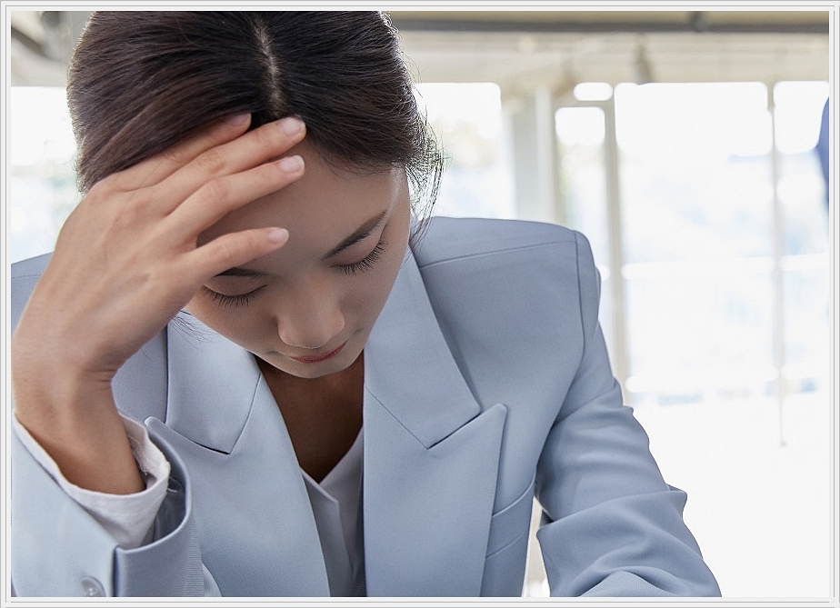 한 손으로 이마를 부여잡고 고개를 숙인 채 다소 힘겨운 표정을 짓고 있는 여성의 모습