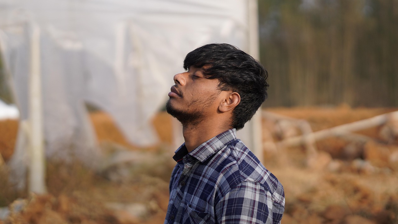 햇살이 따사롭게 비추는 들판을 배경으로, 체크무늬 셔츠를 입은 청년이 눈을 감고 고개를 약간 뒤로 젖힌 채 조용한 호흡을 느끼는 듯한 순간을 포착한 장면이다. 인물의 표정은 평온하면서도 몰입된 상태를 암시하며, 얼굴에 드리운 빛이 따뜻한 분위기를 더한다. 배경은 살짝 흐릿하게 처리되어 있어 초점이 온전히 인물에게 맞춰지며, 인물의 내면 상태에 시선을 집중시킨다. 전체적으로 고요한 명상이나 감정의 침잠을 표현하는 사진.