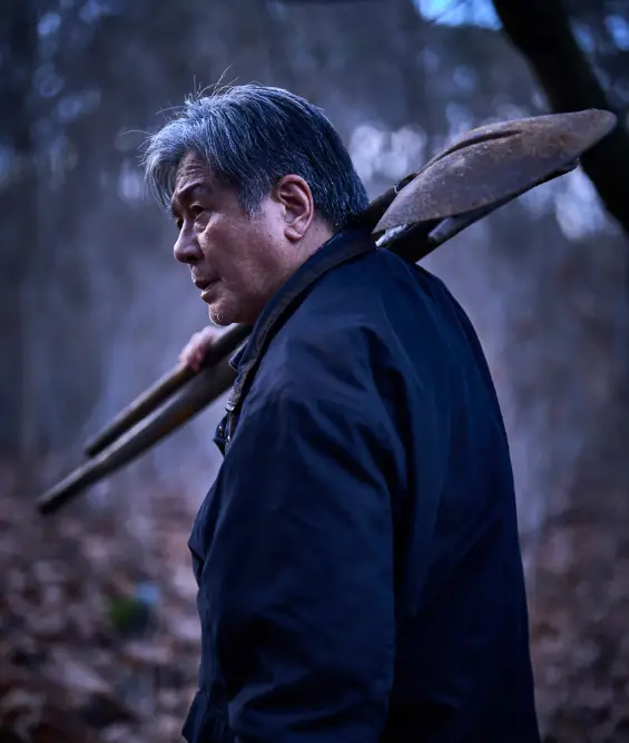 파묘 - 산속 파란옷을 입고 오른쪽 어깨에 삽과 곡괭이를 얹은채 옆으로 서있는 모습