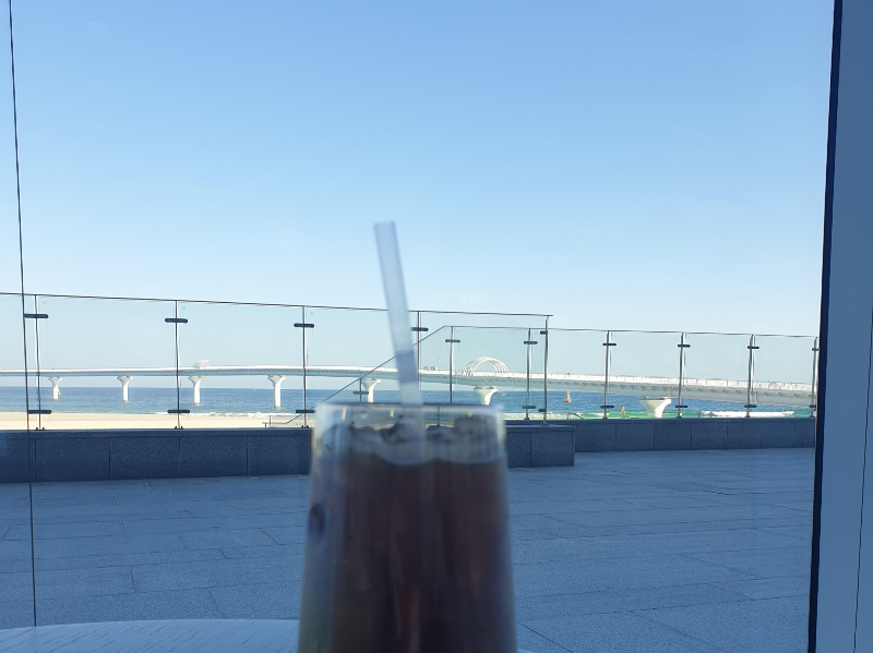 유리창 밖으로 바다와 해변 그리고 다리가 보인다. 그 앞에 아이스 아메리카노