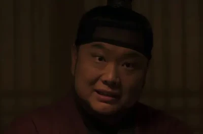 어느 한옥 실내를 배경으로 머리에 상투를 쓰고 붉은 한복을 입고 앉아 있는 드라마 연인의 끗쇠 캐릭터