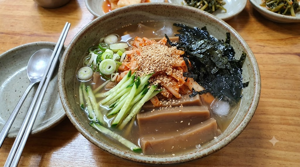 나무 테이블 위에 놓인 시원한 도토리 묵사발 한 그릇의 클로즈업 사진입니다. 도토리묵, 오이채, 다진 김치, 대파, 김가루가 국물과 얼음 위에 넉넉히 올려져 있고, 깨가 뿌려져 있습니다. 옆에는 숟가락과 젓가락, 그리고 다른 반찬들이 보입니다.