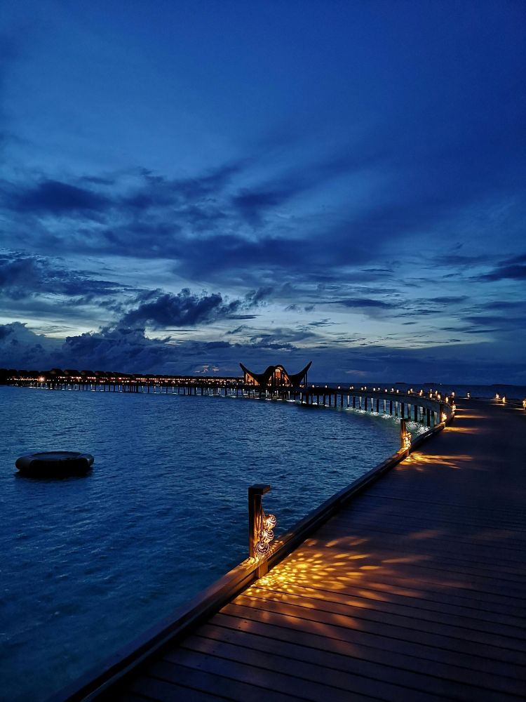 황혼이 내려앉은 몰디브 바다 위 조명으로 밝혀진 곡선형 목조 산책로와 리조트 야경 풍경