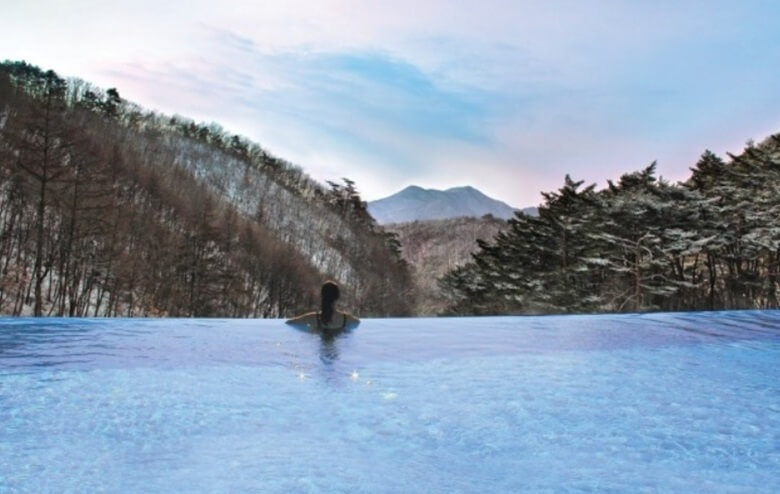 수영장 크기 규모의 야외 온천엔 몸을 담그고 눈덮힌 소나무 숲을 바라보고 있는 한여성의 뒷모습