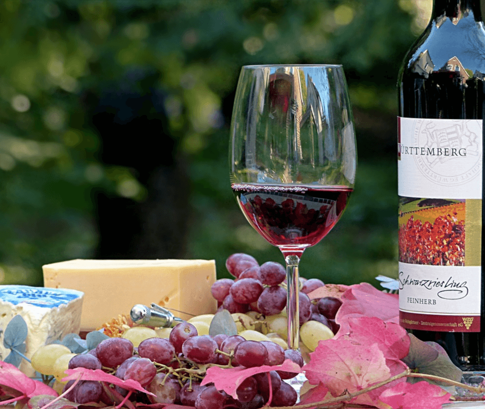 탁자 위에 거봉과 레드와인이 담긴 유리잔, 치즈덩이가 있고 오른 쪽에는 레드와인 병이 놓여있다.