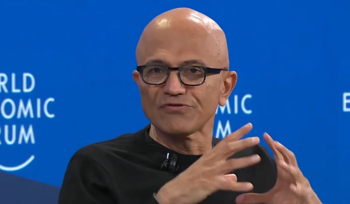 대머리에 안경을 쓴 Microsoft CEO 사티아 나델라가 검은색 터틀넥을 입고 World Economic Forum 무대에서 손동작을 하며 연설하고 있다. 배경은 파란색이며 WORLD ECONOMIC FORUM 문구가 흰색으로 보인다. 나델라는 진지한 표정으로 청중을 향해 말하고 있으며, 전경에는 청중의 뒷모습이 보인다. 전체적으로 고위급 기술 콘퍼런스의 분위기를 전달한다.