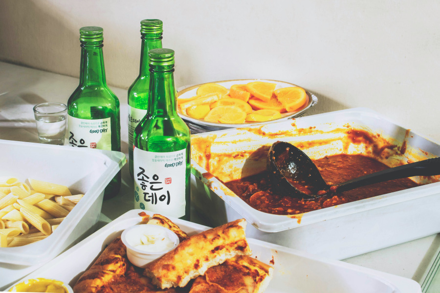 테이블 위에 소주 3병과 피자와 스파게티 면과 국물을 담은 네모난 플라스틱 통과 레몬을 담은 동그란 접시를 놓아두고 찍은 사진