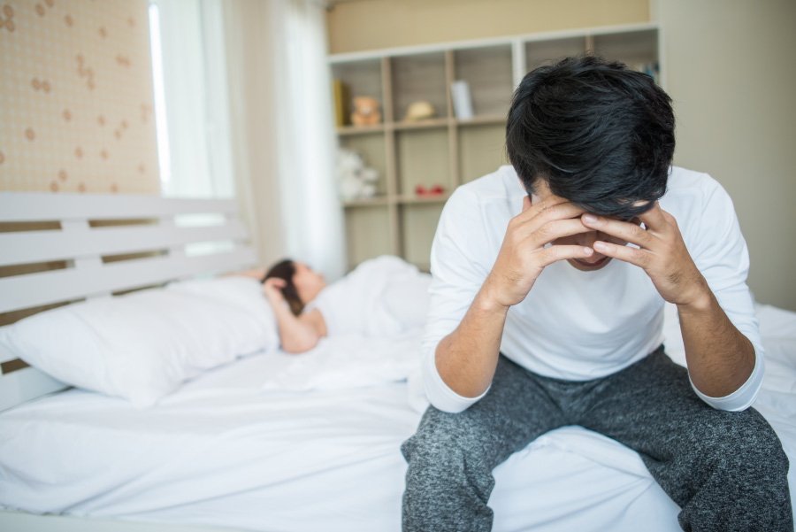 발기부전을 앓고 있는 남성이 침대에 앉아서 양쪽 손으로 머리를 감싸쥐고 있고, 여성은 침대에 누워서 자고 있는 것을 찍은 사진