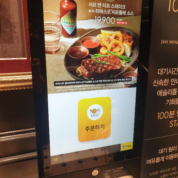 노란 주문하기 버튼이 있는 키오스크. 상단에는 서프 앤 터프 스테이크 with 티바스코 치포틀레 소스라는 광고가 보인다.