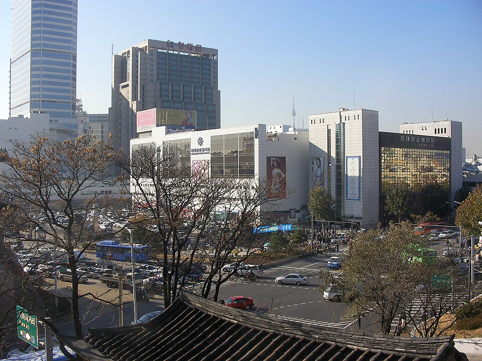 기와지붕 너머로 동대문 일대의 쇼핑몰 건물과 대로가 내려다보이는 맑은 날 풍경