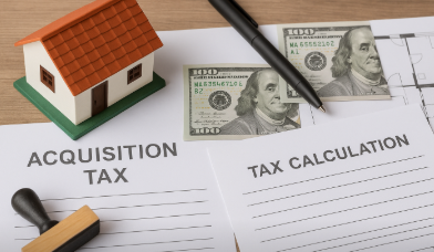 나무 책상 위에 농가주택 모형, 취득세 관련 서류, 도장, 펜, 달러 지폐가 가지런히 놓인 장면