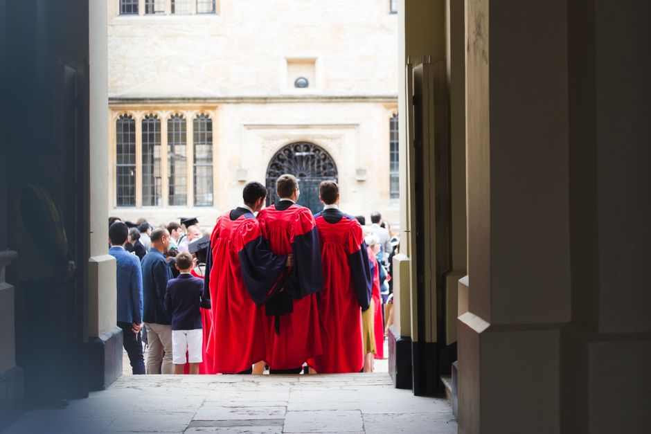 Back view unrecognizable male students wearing red cloaks bonding and standing on crowded square during graduation ceremony in historic university