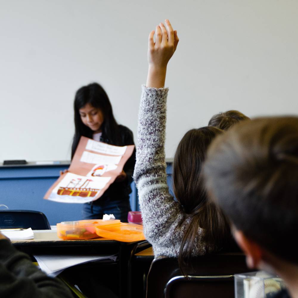 교실에 여러 명의 학생이 의자에 앉아 있고 회색 스웨터를 입은 학생은 한쪽 손을 들고 있다