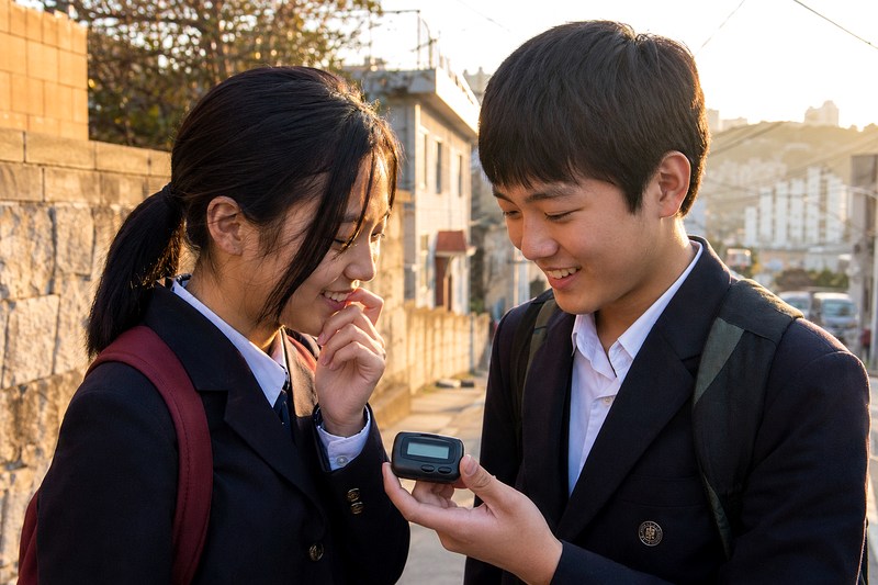 교복을 입은 남학생과 여학생이 골목에서 작은 삐삐 화면을 함께 바라보며 웃는 모습