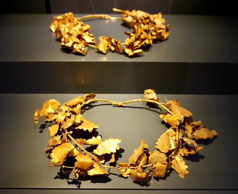 두 개의 금색 월계관이 진열되어 있다. 각각 나뭇잎 모양의 금속 조각들이 얽혀 있으며, 실제 식물처럼 자연스러운 곡선과 구조를 가지고 있다.