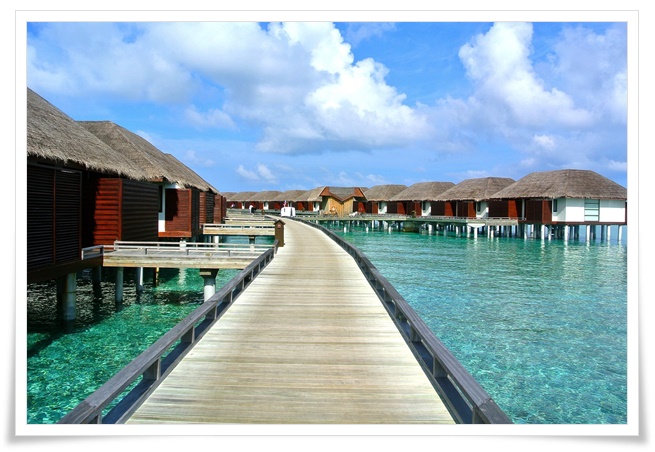 에메랄드빛 바다와 초가지붕의 수상빌라가 조화를 이루는 해변휴양지의 모습을 잘 보여주고 있음. 긴 데크 다리로여러채의 수상빌라들이 연결되어 바다물 위의 빌라 리조트가 매우 고급스럽게 보임