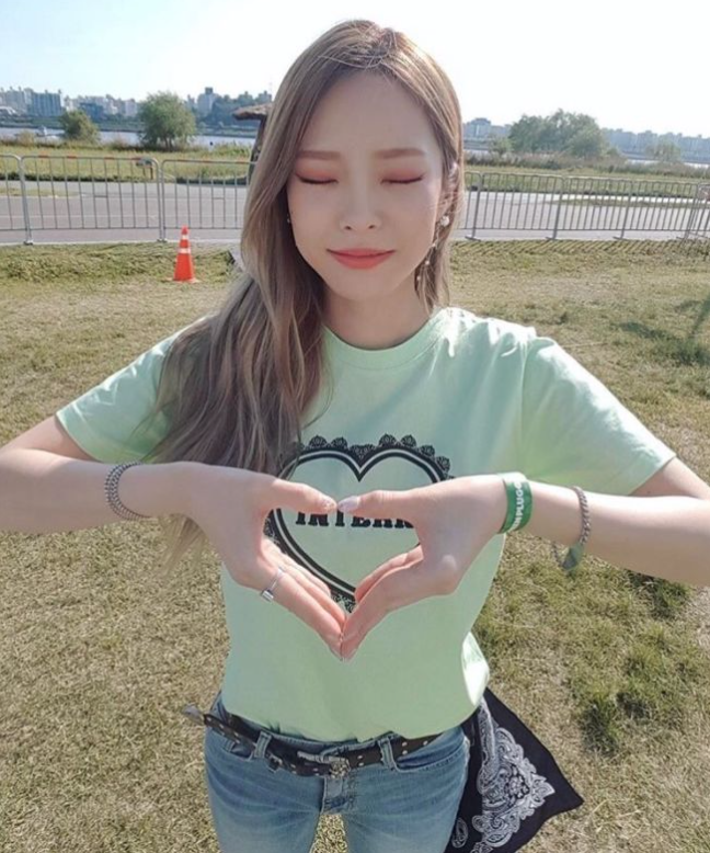 헤이즈가-햇살-좋은-날-청바지와-연두색-반팔티를-입고-손으로-하트를-그리며-눈을-감고-미소짓고-있다.