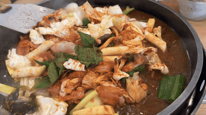 철판 위에서 닭고기, 떡, 채소가 빨간 양념과 함께 보글보글 끓고 있는 송정 닭갈비. 깻잎과 양배추가 서서히 양념에 물들어가고 있다.