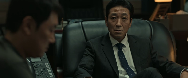 검은 양복을 입고 쇼파에 앉아 있는 독전2의 청장 역을 맡은 배우 최광일