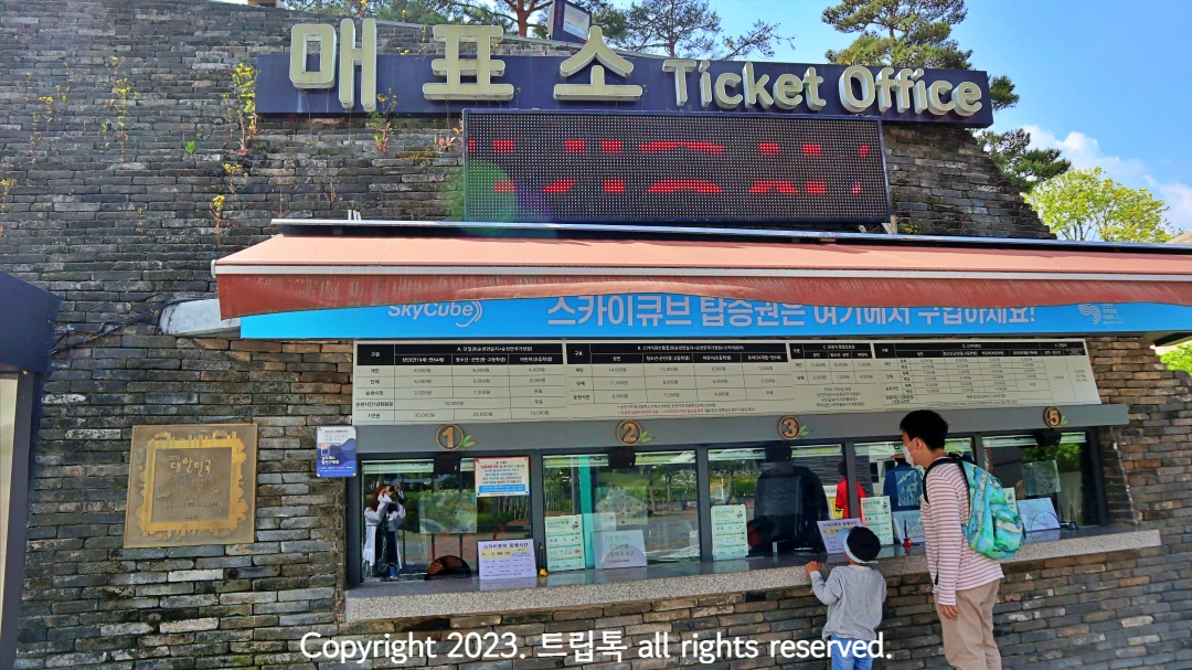 파란색 간판에 하얀 글씨로 매표소라고 적힌 벽돌 건물이 있고 5개 창구에서 매표를 하고 있다. 3번 창구에서 두 사람이 입장권을 산다.