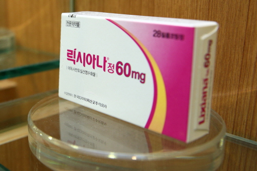 유리-선반-위에-진열된-릭시아나정-60mg-포장-박스의-측면-사진-박스는-흰색-바탕에-분홍색과-노란색-곡선-디자인이-돋보이며-전면에는-진한-빨간색과-보라색-글씨로-제품명과-용량-복용-정보가-표기됨-상단에는-전문의약품-표시가-작게-인쇄되어-있고-조명이-반사된-투명한-원형-유리-진열대에-깔끔하게-배치되어-있는-구성