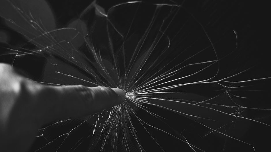 Black and white close-up of a finger touching a cracked glass screen, creating a dramatic effect.