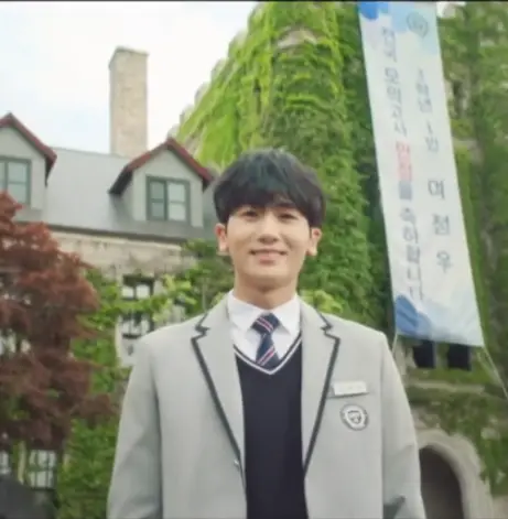 닥터슬럼프- 건물에 초록색 나뭇잎들이 감싸져있고 그앞 회색 쟈켓 남색조끼 흰셔츠 남색 넥타이를 메고 정면을 보며 웃고있는 박형식