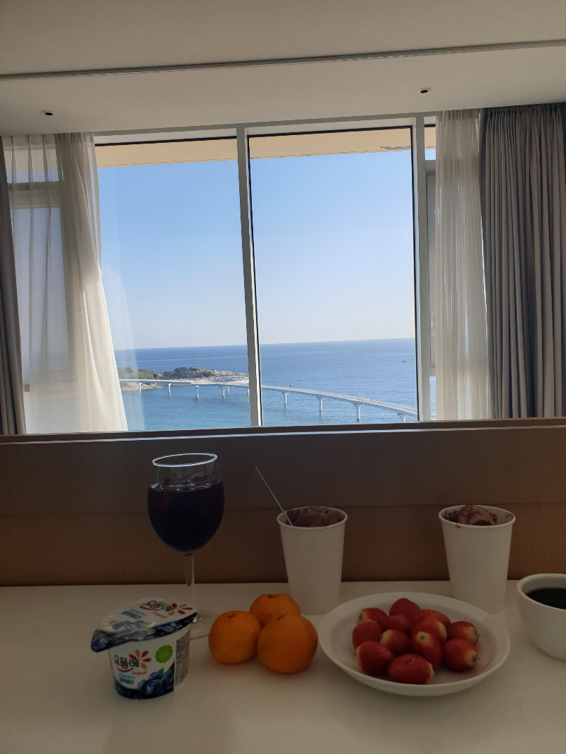 바테이블 위에 커피와 과일 그리고 요거트와 커피가 있다. 창문 너머로는 바다가 보인다.