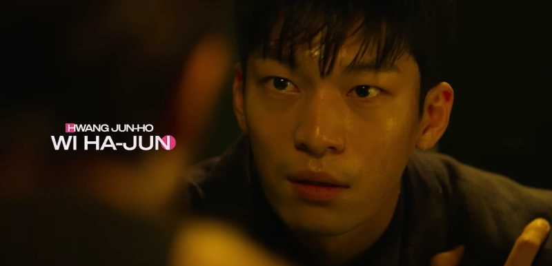 어두운 실내를 배경으로 누군가를 바라보고 있는 오징어 게임의 황준호를 연기하는 배우 위하준