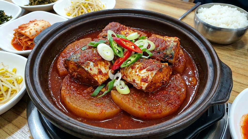검은색 뚝배기에 담긴 매콤한 고등어조림. 양념이 잘 밴 두툼한 무와 고등어 토막 위에 송송 썬 대파와 고추가 고명으로 올라가 있다. 주변에는 공기밥과 김치, 콩나물무침 등의 밑반찬이 함께 차려져 있다.
