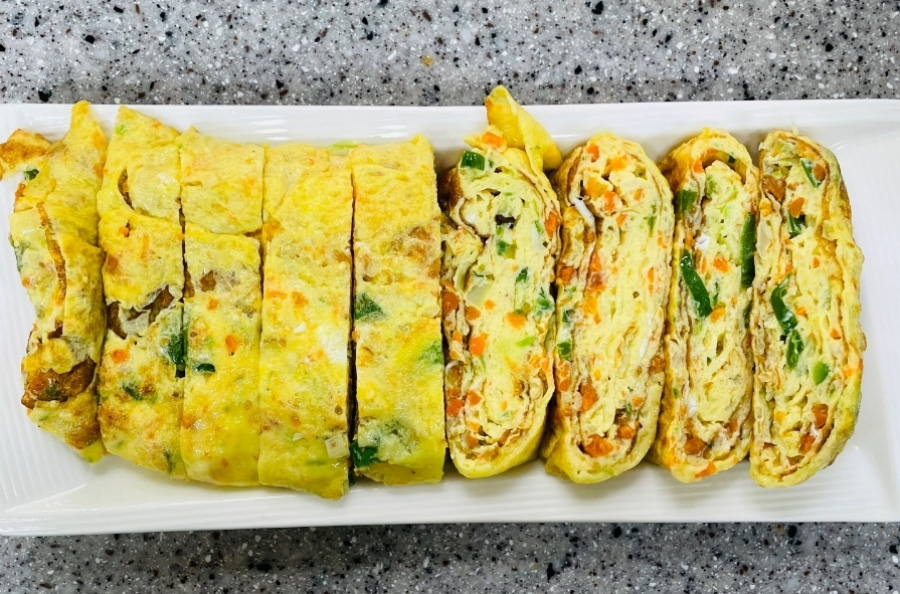 각종 야채와 고기를 다져서 계란을 푼 물에 넣고 후라이팬에서 익혀서 만든 달걀야채말이를 희고 네모난 그릇에 가지런히 담아서 바닥에 놓아두고 위에서 찍은 사진