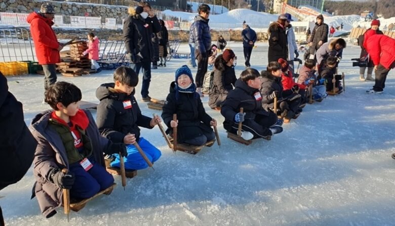 아이들이 한줄로 늘어서서 얼음위에 썰매를 타고 대기하고 있고 그 뒤에서는 부모님들이 출발 신호를 기다리는 듯 준비하고 있는 모습