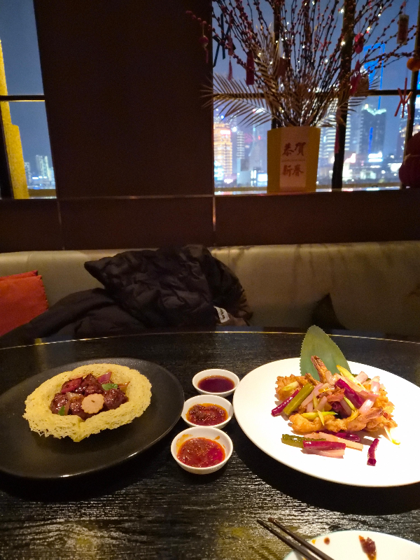 창 밖으로 어두워진 하늘 그리고 조명으로 밝아진 빌딩. 앞에는 페퍼 비프와 크리스피 새우가 올려져 있다.